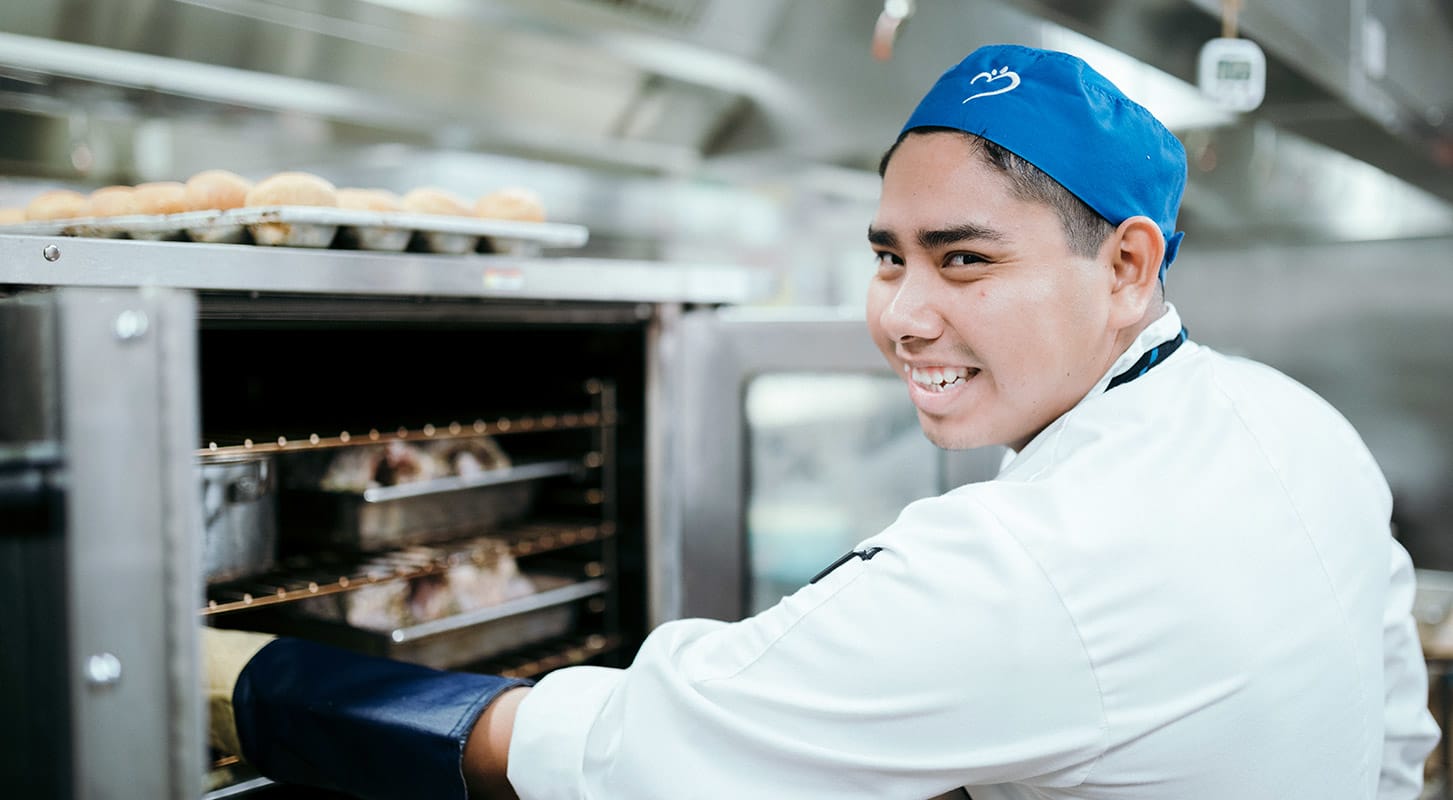 A chef wearing a white shirt and TouchPoint blue hat smiles as he pulls warm bread from the oven.